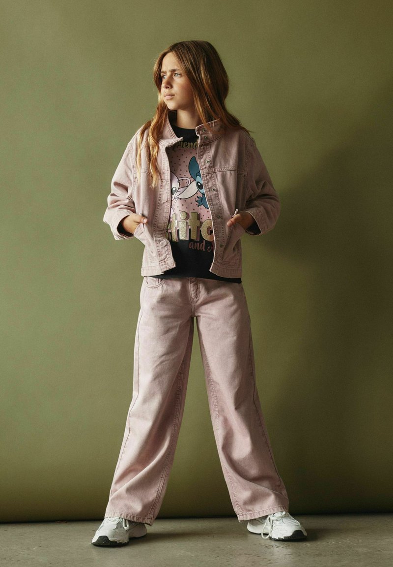 Fille aux longs cheveux portant une veste rose clair et un pantalon, un t-shirt noir à motif et des baskets blanches, debout contre un mur vert olive.
