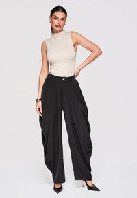 Femme debout, main sur la hanche, portant un haut beige sans manches et un pantalon noir plissé à jambes larges, avec des talons hauts noirs.