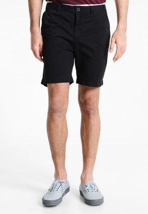 Shorts en coton noir à coupe droite, avec une fermeture à bouton à l'avant et des poches latérales. Portés avec des baskets gris clair.