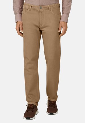 Mann trägt hellbraune gerade geschnittene Hose mit Knopf und Reißverschluss, kombiniert mit braunen Sneakers und einem langärmligen Hemd.