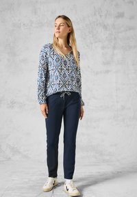 Langarmbluse mit Muster in Blau und Weiß, kombiniert mit navyfarbener Hose und weißen Sneakern. Die Bluse hat einen V-Ausschnitt und Knopfdetails.