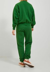 Sudadera verde hecha de tejido suave, con un corte holgado, puños acanalados y pantalones ajustados a juego. Las zapatillas son de un tono beige claro con detalles en negro.