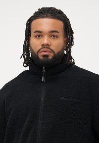 Mand med mellemstore dreadlocks og skæg iført en sort lynlås fleecejakke, vendt fremad mod en ensartet baggrund.