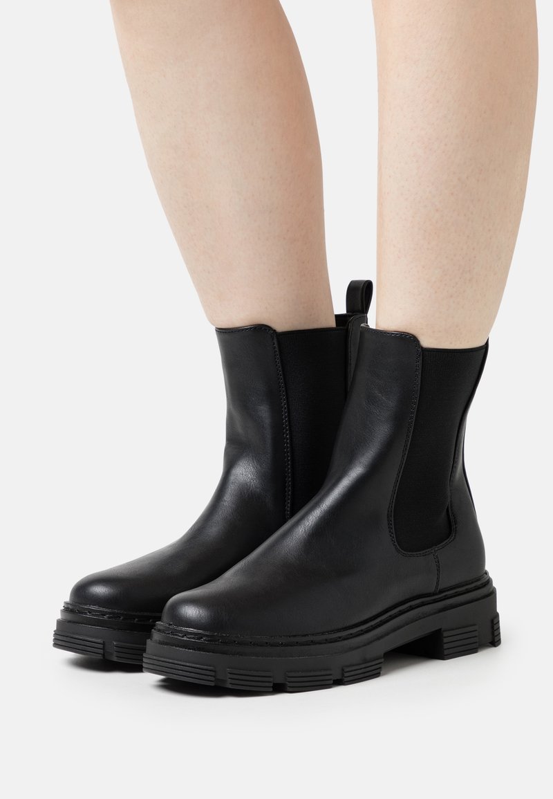 Jambes portant des bottines en cuir noir avec des semelles épaisses en caoutchouc et des panneaux latéraux élastiques sur un fond clair uni.