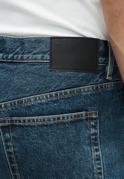 Πίσω τσέπη και μέση μπλε τζιν με μαύρη ορθογώνια δερμάτινη ετικέτα επωνυμίας πάνω από την τσέπη, φορεμένα με λευκό τοπ.