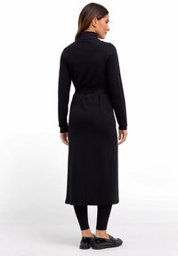 Kobieta w długiej czarnej sukience-płaszczu z wysokim kołnierzem i paskiem, czarnych legginsach i czarnych loafersach, stojąca tyłem na białym tle.