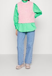 Henkilö, joka on pukeutunut kirkkaan vihreään pitkähihaiseen paitaan, tekstuurilla varustettuun vaaleanpunaiseen hihattomaan liiviin, vaaleansinisiin farkkuihin ja beigeihin lenkkareihin seisoo valkoista seinää vasten.