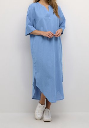 Femme portant une robe midi ample bleu clair à manches mi-longues et des baskets blanches, debout devant un fond blanc uni.
