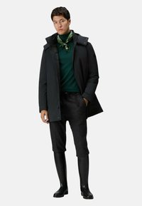 Zwarte waterdichte jas, groene coltrui, groen patroon sjaal, grijze geklede broek en zwarte rubberen laarzen tot de knie.