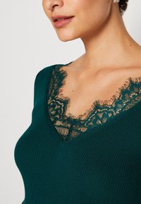 Femme portant un haut côtelé turquoise à encolure en V ornée d'une finition en dentelle noire à motifs floraux.