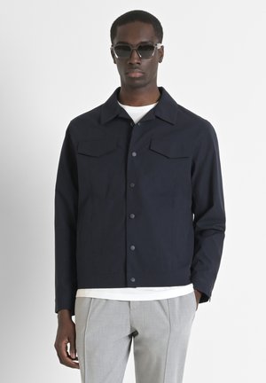 Chaqueta navy con dos bolsillos en el pecho, cierre de botones y cuello plano. Combinada con pantalones gris claro y una camisa blanca, que presenta una textura suave.