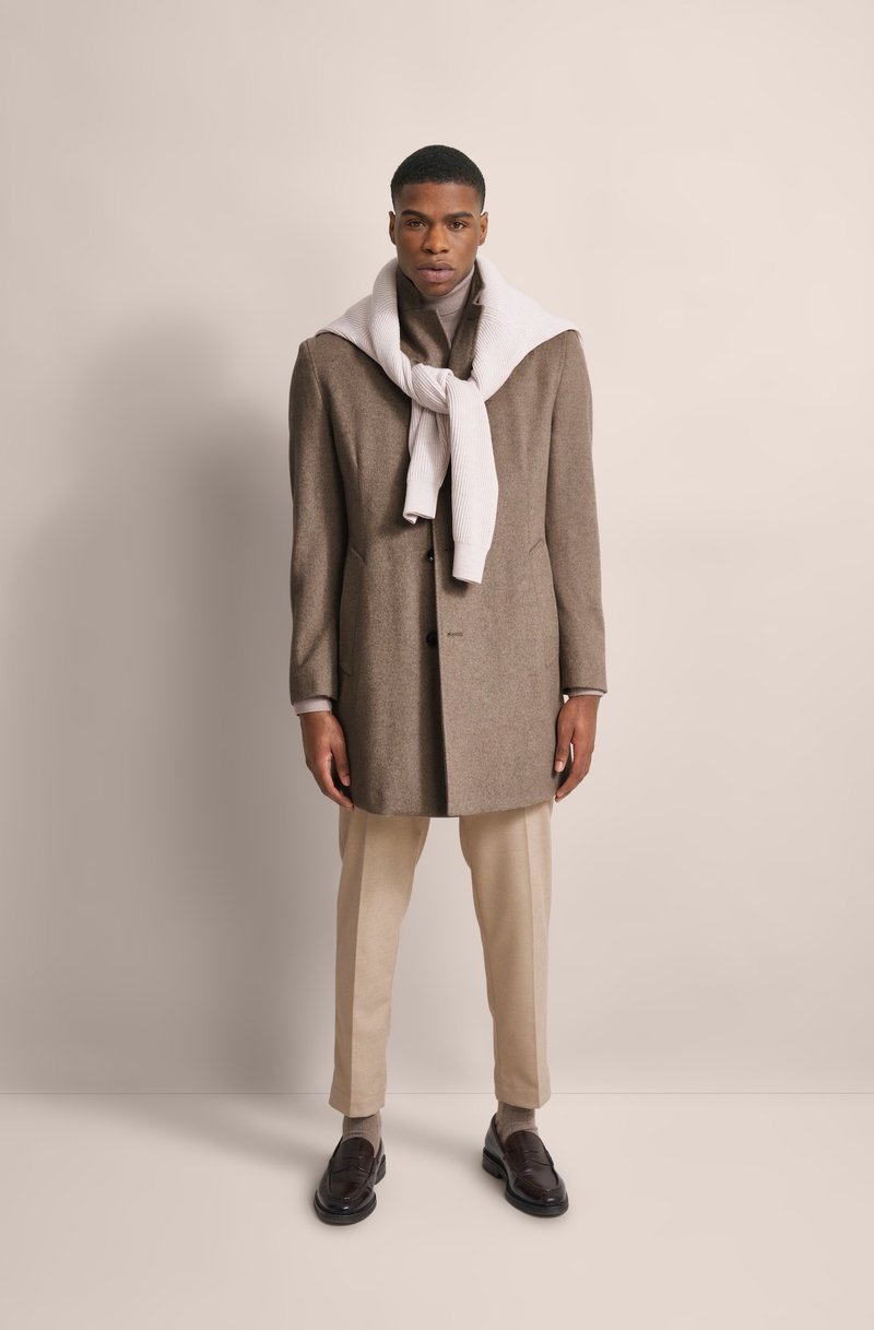 Cappotto di lana marrone con vestibilità sartoriale, chiusura a bottoni e tasche laterali doppie. Sciarpa bianca a coste drappeggiata attorno al collo, pantaloni beige e scarpe scure.
