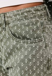 Jeans de denim verde con patrones bordados en blanco, mostrando un primer plano de la cintura y los bolsillos, destacando un acabado texturizado.
