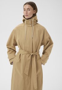 Žena v béžovém plášti s kapucí a páskem, s rukama v kapse, stojící na jednobarevném světlém pozadí.