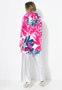 Květinový kabát s kapucí v živých odstínech růžové, bílé a tyrkysové; má dlouhé rukávy a volný střih, který je doplněn lesklými stříbrnými kalhotami s širokými nohavicemi.