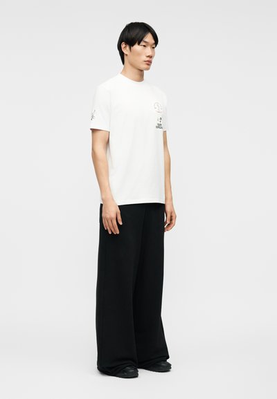 Άνδρας στέκεται φορώντας ένα λευκό μπλουζάκι με σχέδιο και φαρδιές μαύρες παντελόνες, μπροστά σε απλό ανοιχτό γκρι φόντο.