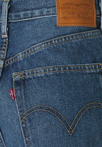 Siniset denim-farkut, joissa on näkyvät ompeleet, nahkalenkki vyötäröllä ja punainen Levi's-logo takataskussa. Ominaisuutena klassinen niittisuunnittelu.
