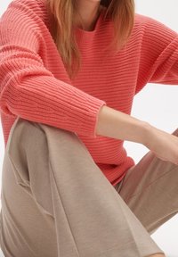 Sweter o koralowej teksturze, wykonany z dzianiny, z szerokimi rękawami, zestawiony z jasnobeżowymi spodniami typu wide-leg, prezentujący miękki materiał i luźny krój.