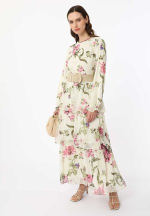 Donna che indossa un lungo vestito bianco a fiori con motivi rosa e verdi, una cintura beige intrecciata, tiene una borsa beige e indossa sandali con cinturini.