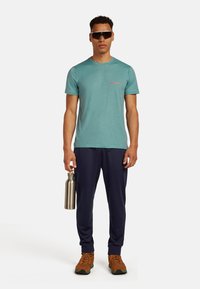 Lys teal kortærmet t-shirt med lille rød logo, marineblå sweats, holder en sølvvandflaske, brune vandresko.