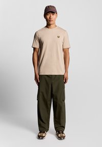 T-shirt di cotone beige con un piccolo logo di farfalla nera, abbinato a pantaloni cargo verdi e sneaker con motivo marrone. Cappellino grigio in testa.