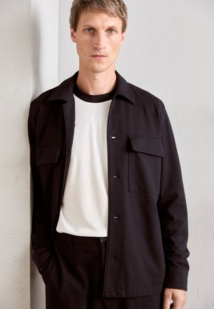 Man with light brown hair wearing a black jacket over a white ribbed shirt, standing against a light-colored textured wall.