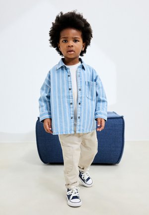 Lille barn går indendørs iført en blåstribet skjorte, beige bukser og marineblå sneakers, med en blå skammel i baggrunden.