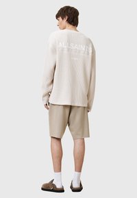 Top de manga larga beige con tejido texturizado y el logo "ALL SAINTS" en la parte posterior, combinado con pantalones cortos beige claros y zapatos marrones.