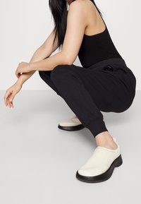 Vita slip-on skor med en slät, glänsande textur och svarta gummisulor. Kombineras med svarta joggarbyxor och en åtsittande svart topp.