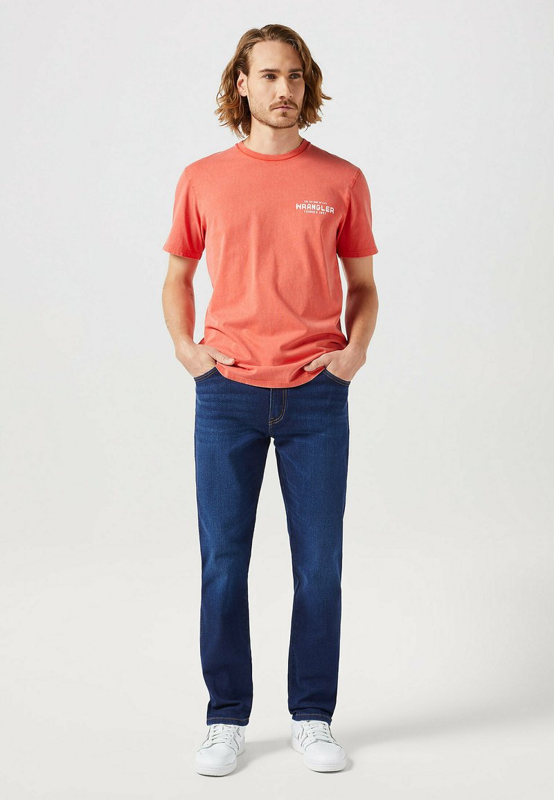 Korallowy t-shirt z krótkim rękawem z logo "Wrangler", w połączeniu z ciemnoniebieskimi dżinsami i białymi sneakersami, stojący na jasnym tle.