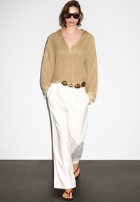 Top oversized a righe beige con scollo a V, pantaloni bianchi a vita alta e infradito rosse. Una cintura con dettagli dorati completa l'outfit.