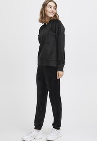 Tuta in velour nera con felpa a cappuccio e tasca frontale, pantaloni slim-fit. Abbinata a scarpe da ginnastica bianche. Texture liscia.