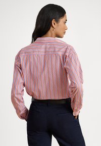 Sieviete ar gariem tumšiem matiem, valkājot sarkanu, baltu un zilu vertikāli svītrainu kreklu, kas ielikts tumši zilos biksēs, stāv ar rokām kabatās.