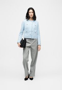 Ženska v svetlo modri teksturirani jakni, sivo hlače s črtami, črni čevlji z visokimi petami, drži črno torbico na enobarvnem ozadju.