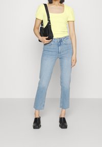 Keltainen röyhelöillä varustettu lyhythihainen paita, korkeavyötäröiset vaaleansiniset farkut, joissa on raikkaat helmat, ja mustat palttinadonnat, joita pidetään mustalla olkalaukulla.