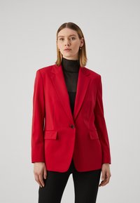 Červený blazer s klopami, jedním knoflíkem, dvěma předními kapsami a hladkou strukturou, nošený přes černý top.