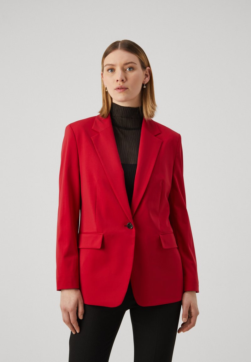 Červený blazer s klopami, jedním knoflíkem, dvěma předními kapsami a hladkou strukturou, nošený přes černý top.