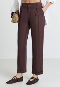 Pantaloni a pieghe marroni con una texture liscia, vita alta e chiusura con un solo bottone. Inclusi risvolti e tasche laterali.