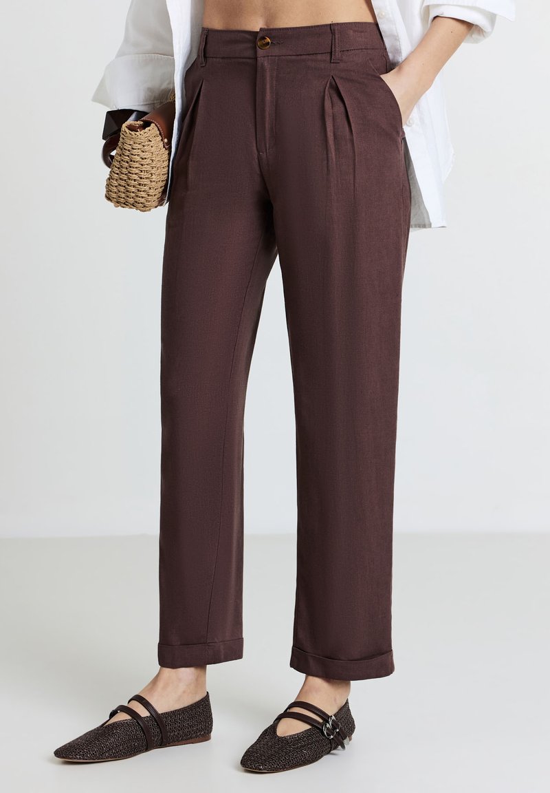 Pantaloni a pieghe marroni con una texture liscia, vita alta e chiusura con un solo bottone. Inclusi risvolti e tasche laterali.
