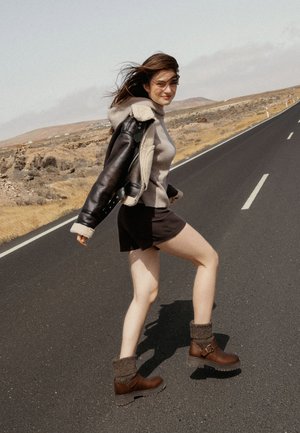 Botas de tobillo marrones con una suela gruesa y puños de punto grises, combinadas con un suéter gris y una chaqueta de cuero negra con forro de borreguito.