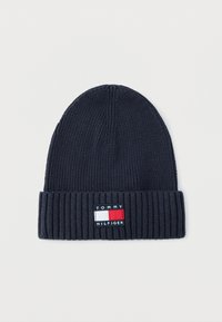 Temno mornarska pletena kapa z rebrastim robom, na katerem je pravokotna etiketa z rdečo, belo in modro znamko. Mehka tekstura, valjaste oblike.