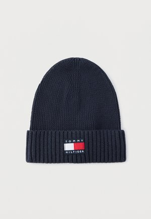 Bonnet en tricot bleu marine foncé avec bord côte, doté d'une étiquette rectangulaire avec un logo rouge, blanc et bleu. Texture douce, forme cylindrique.