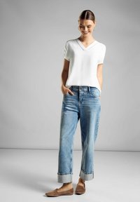 Maglietta bianca a V con maniche corte, jeans dritti azzurri chiari con risvolti, e scarpe slip-on marroni. Texture lisce e vestibilità casual.