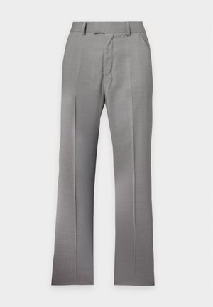 Mann steht mit den Händen in den Taschen grauer Hosen, trägt ein dunkelblaues Poloshirt mit weißer Einfassung und schwarze Anzugschuhe.