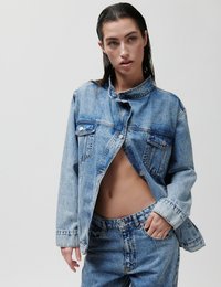 Νεαρή γυναίκα με βρεγμένα μαλλιά που φοράει ένα ανοιχτό ανοιχτό γαλάζιο τζιν μπουφάν και ταιριαστό τζιν ψηλόμεσο παντελόνι, σε ουδέτερο φόντο.