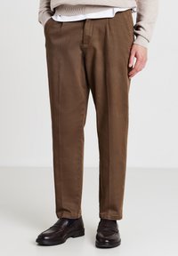 Mann in braunen, gerade geschnittenen Hosen, beigem Pullover, weißem Hemd und dunkelbraunen Lederloafers, der auf einem weißen Boden steht.