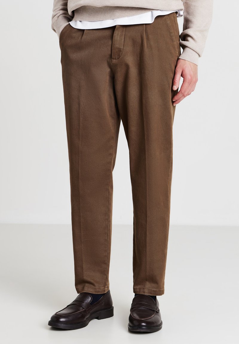 Mann in braunen, gerade geschnittenen Hosen, beigem Pullover, weißem Hemd und dunkelbraunen Lederloafers, der auf einem weißen Boden steht.