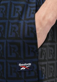 Reebok šorti ar tumši zilu ģeometrisku rakstu un "R" logotipiem, izgatavoti no mīkstas auduma. Ietver zīmola uzšuvi ar sarkaniem, baltiem un ziliem akcentiem.
