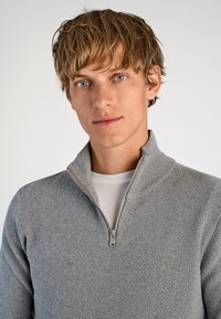 Grauer, mit Reißverschluss versehenen Pullover mit strukturiertem Strickmuster. Verfügt über einen Stehkragen und wird über ein weißes Hemd getragen.