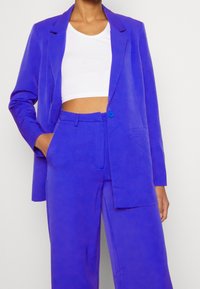 Blazer ajusté violet vif avec un pantalon assorti. Le blazer est doté d'un seul bouton bleu, de revers en pointe et de deux poches avant. Le pantalon présente une coupe décontractée.